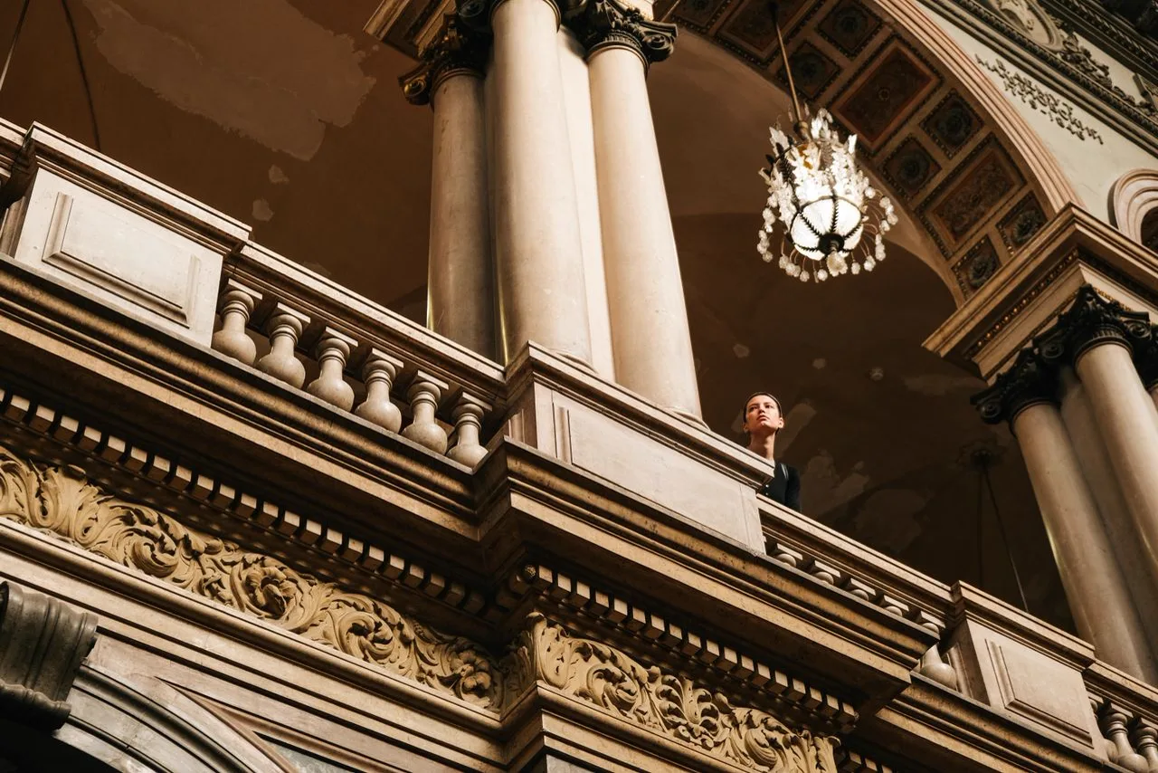 A person looking out from an ornate stone balcony beneath columns and a chandelier