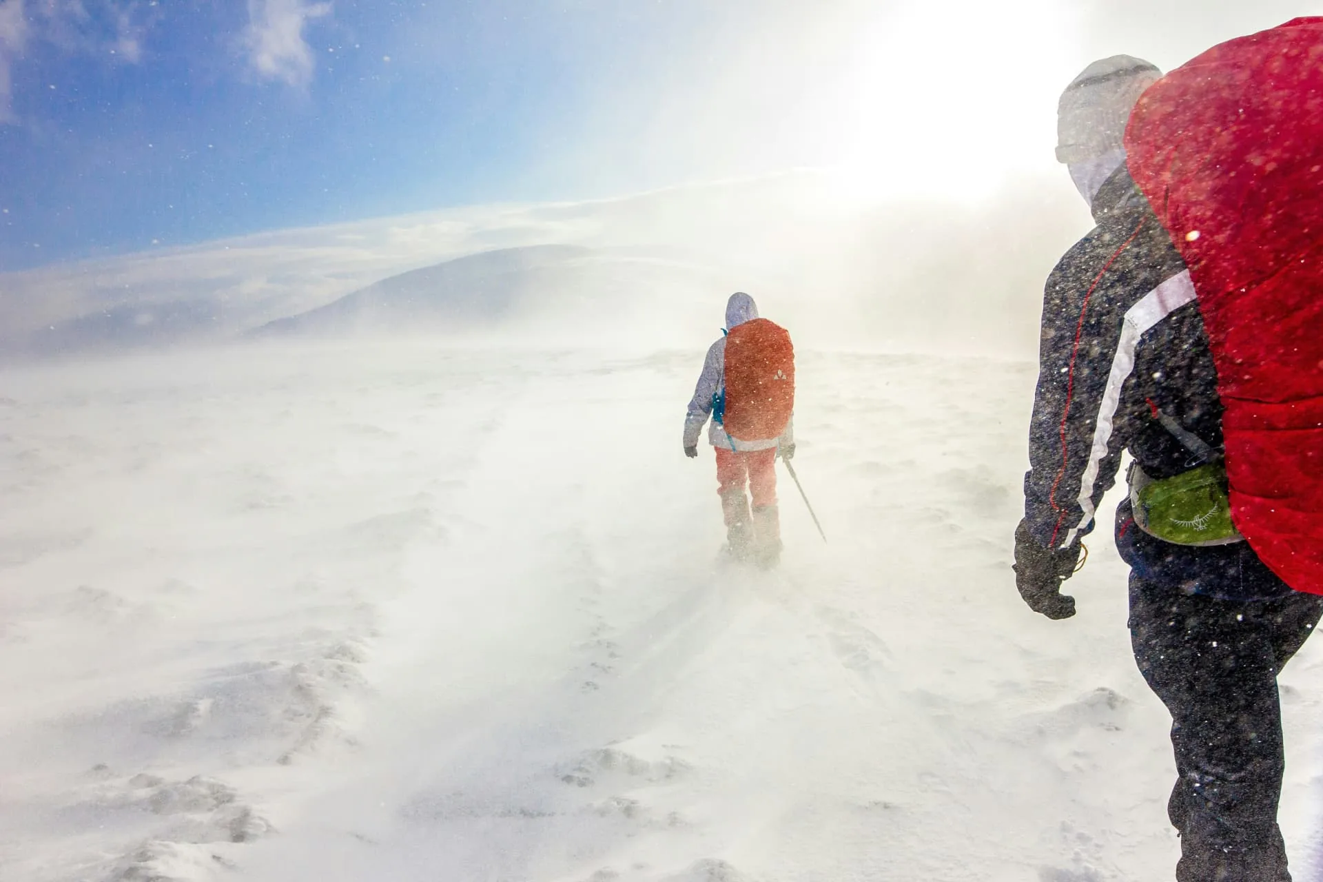 Two hikers carrying backpacks through windblown snow on a mountain