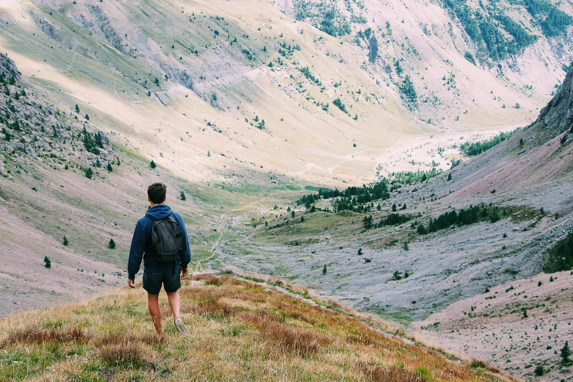 A hiker with a backpack walking down a grassy ridge into a wide mountain valley
