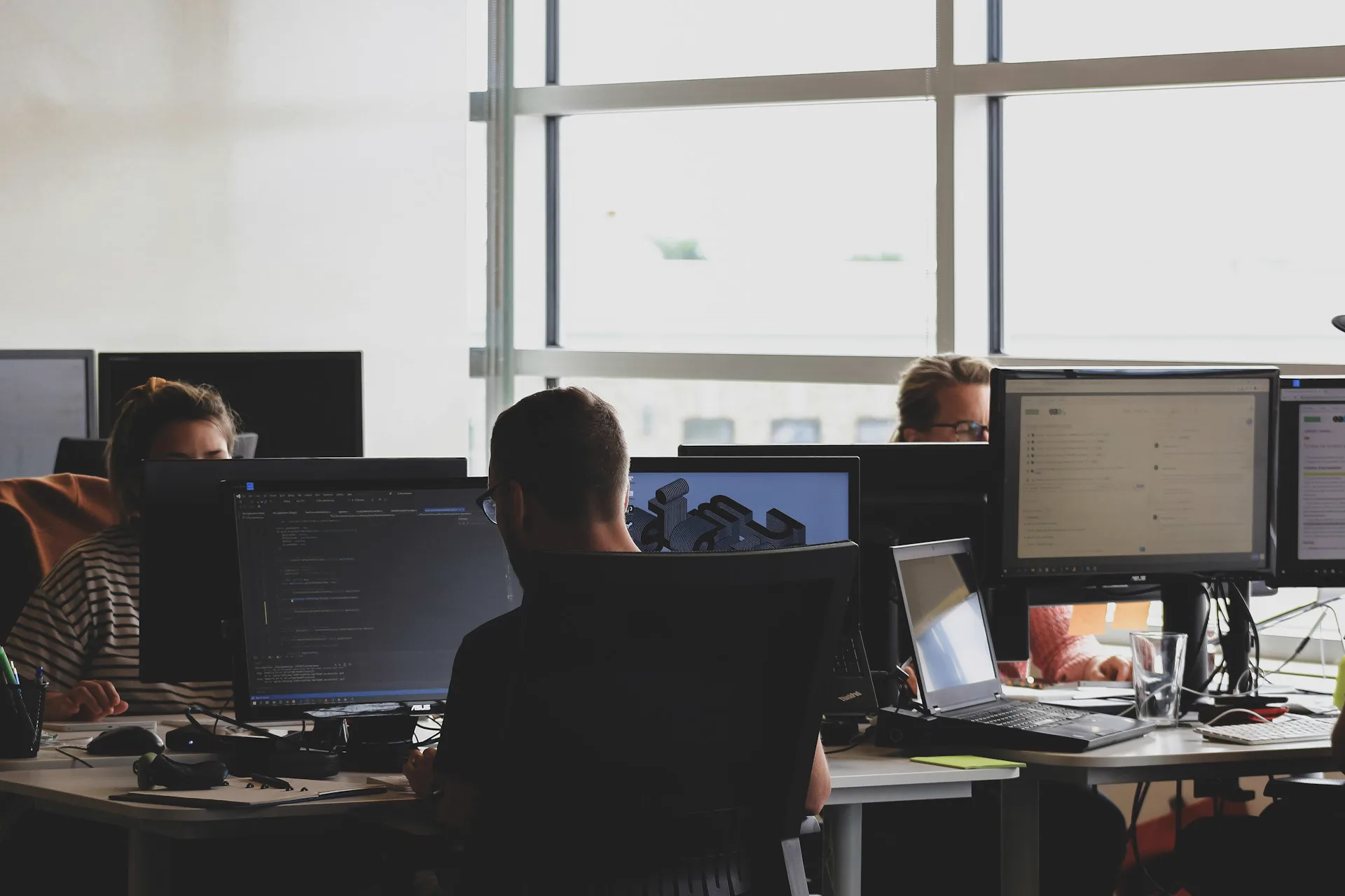 People at shared desks, each focused on their own computer screens, in front of large windows